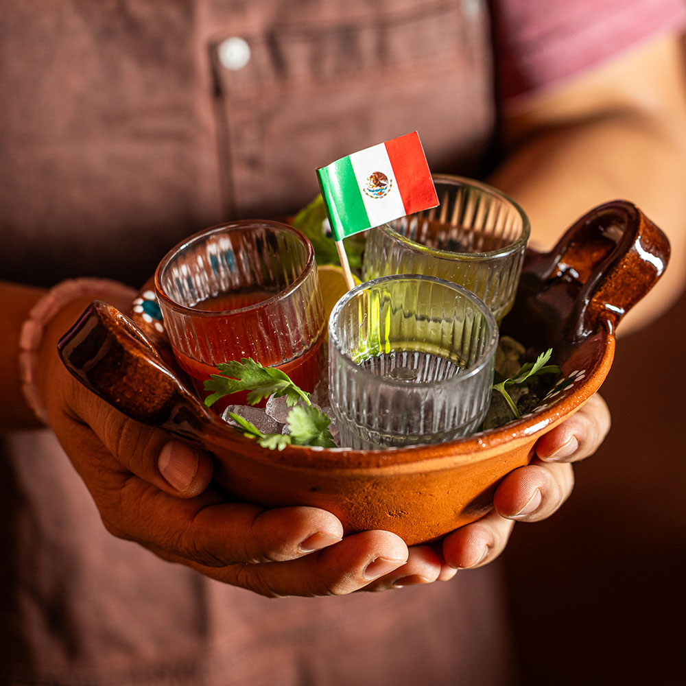 Man holding bowl with 3 small beverages and Mexican flag